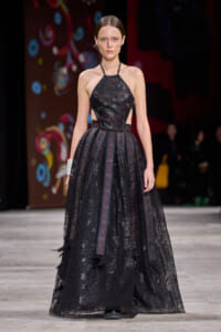 Model on a runway wearing a black lace halter-neck gown with a full sheer skirt on a fashion show stage.