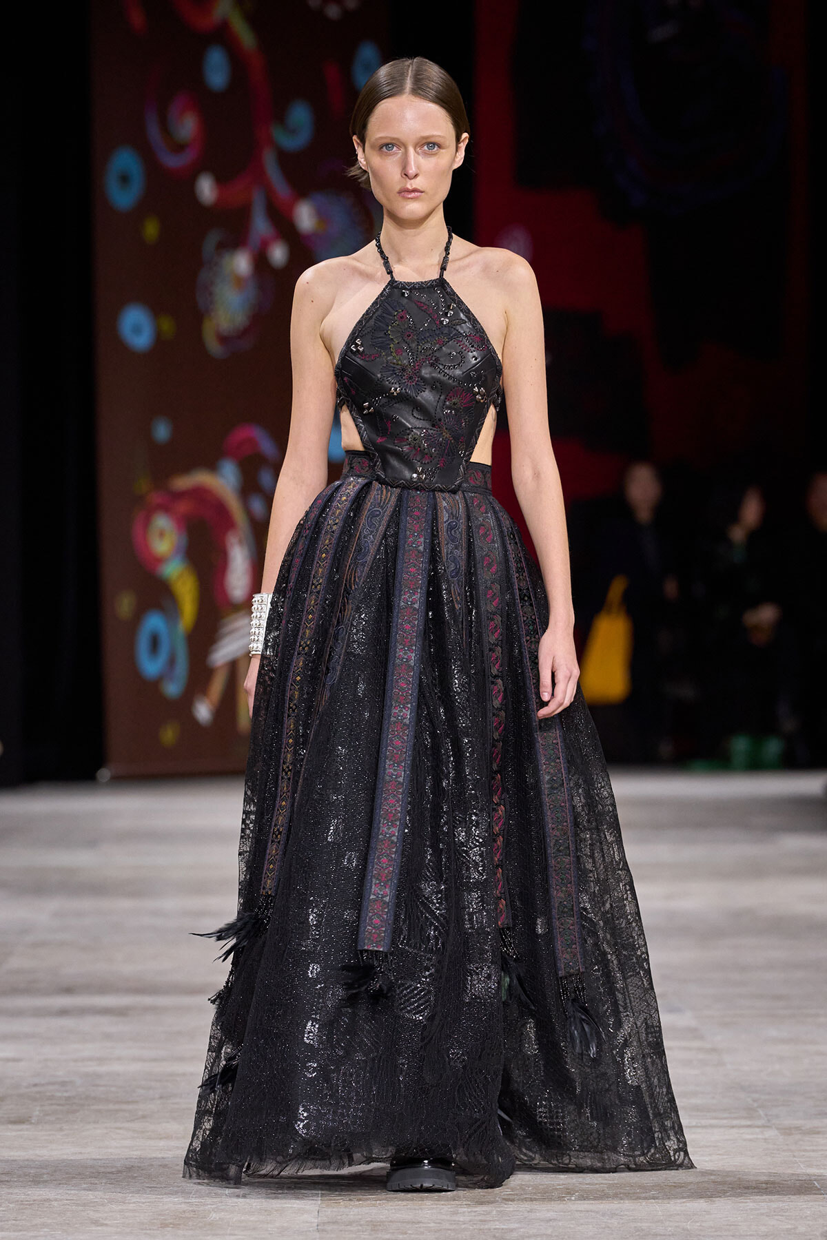 Model on a runway wearing a black lace halter-neck gown with a full sheer skirt on a fashion show stage.