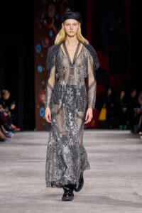 Model on runway wearing a long, sheer silver gown with a snakeskin-like pattern and black boots, paired with a black cap.