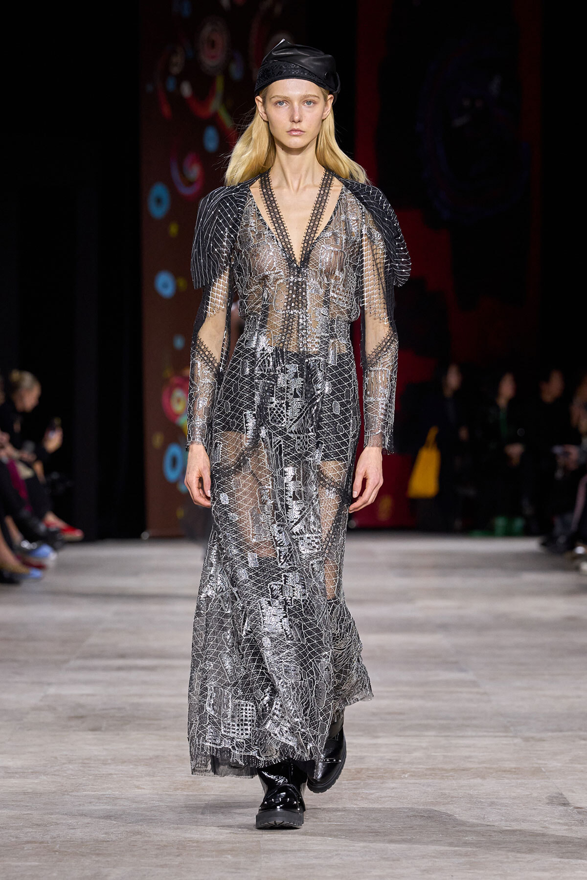 Model on runway wearing a long, sheer silver gown with a snakeskin-like pattern and black boots, paired with a black cap.
