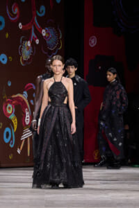 Model wearing a floor-length black lace gown with side cutouts, walking a fashion runway against a colorful painted backdrop.