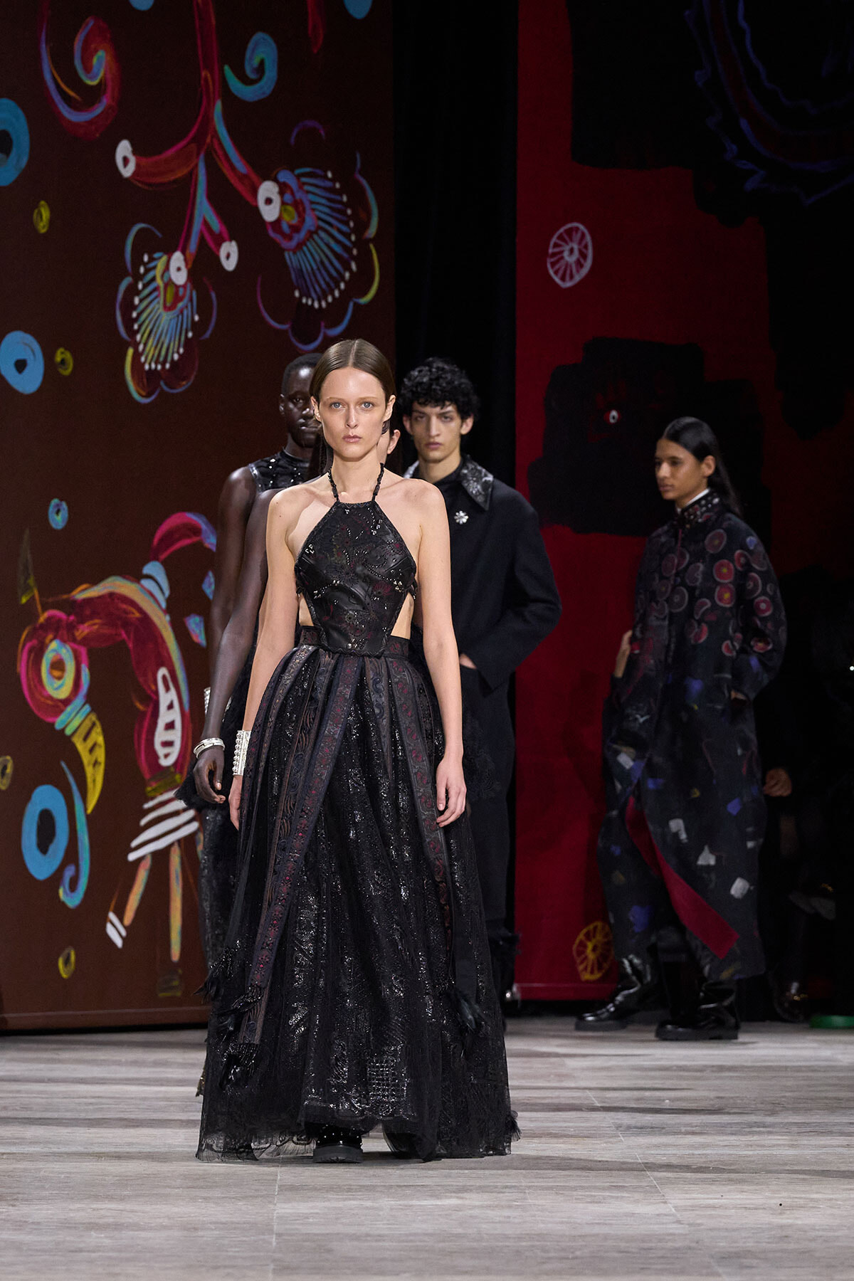 Model wearing a floor-length black lace gown with side cutouts, walking a fashion runway against a colorful painted backdrop.