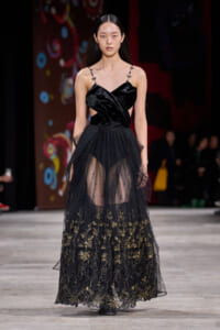 Model walks runway in a black satin gown with a structured bodice and sheer, gold-embroidered lace skirt.