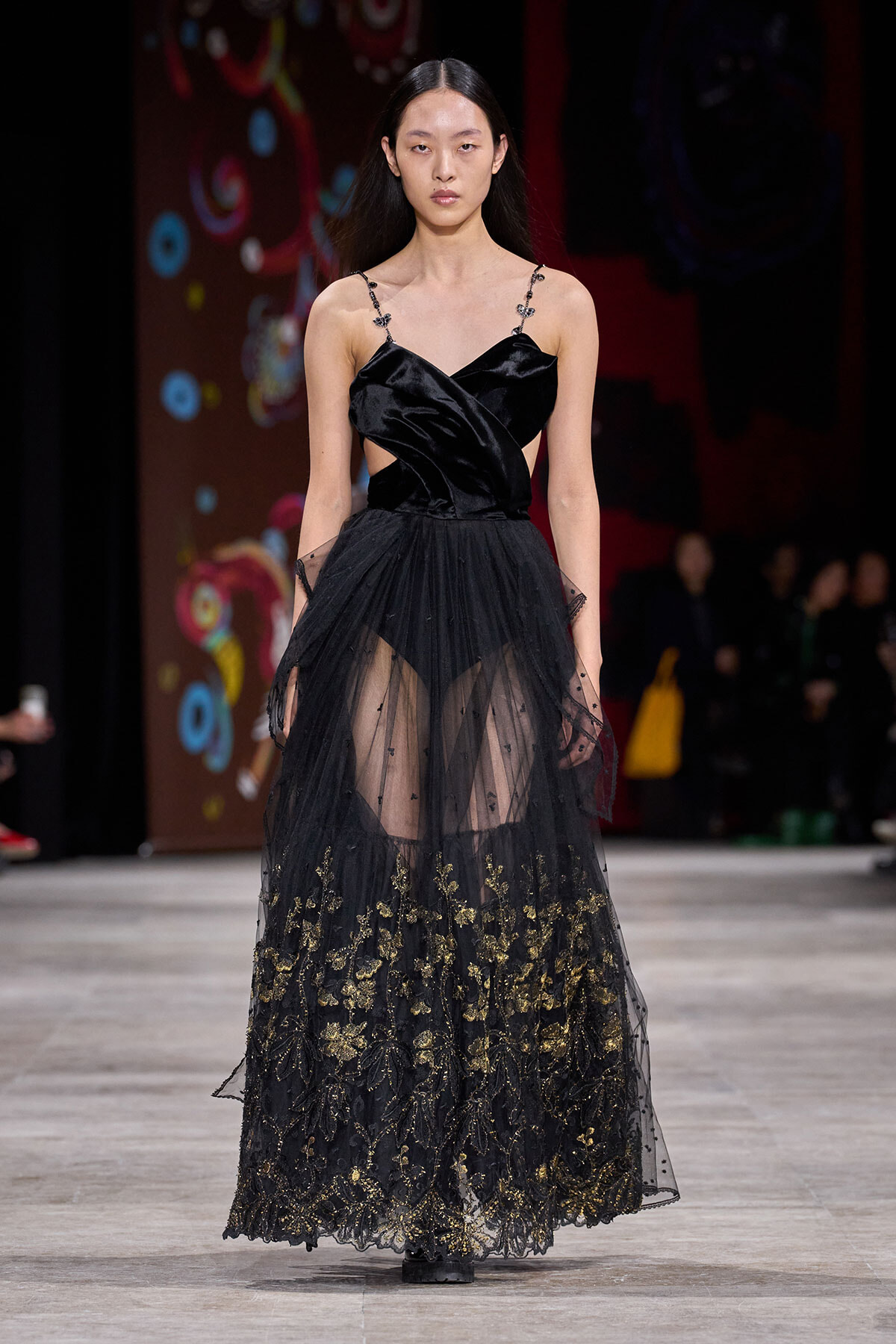 Model walks runway in a black satin gown with a structured bodice and sheer, gold-embroidered lace skirt.