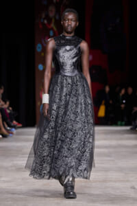 Model walks the runway in a black lace evening gown with a metallic embroidered bodice, full lace skirt, and a wide silver cuff bracelet on her left wrist.