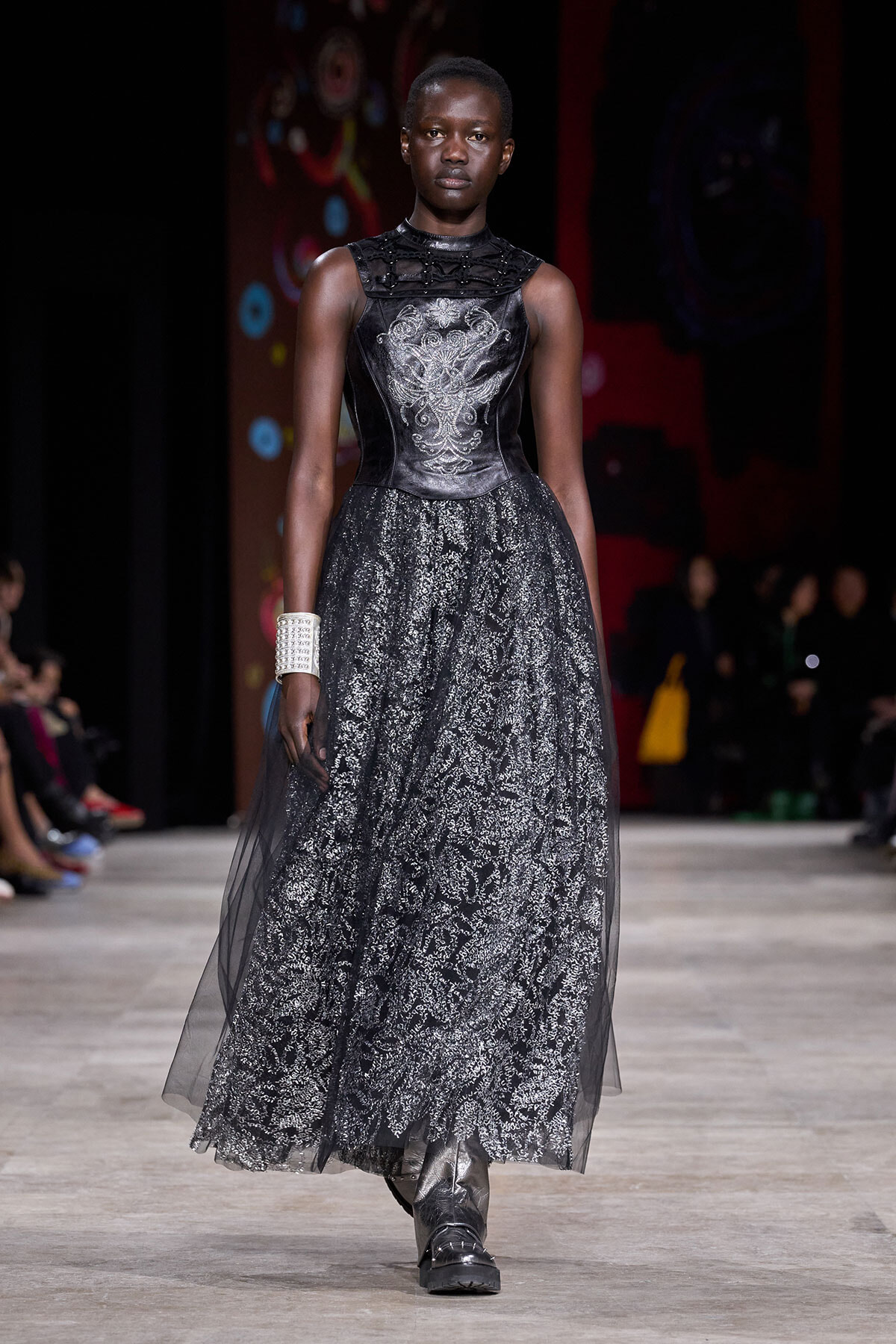 Model walks the runway in a black lace evening gown with a metallic embroidered bodice, full lace skirt, and a wide silver cuff bracelet on her left wrist.