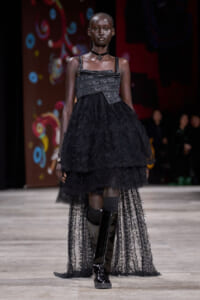 Black-clad female model walking a runway in a layered lace dress with a metallic corset-style bodice, choker, and tall shiny boots; colorful stage backdrop in background.