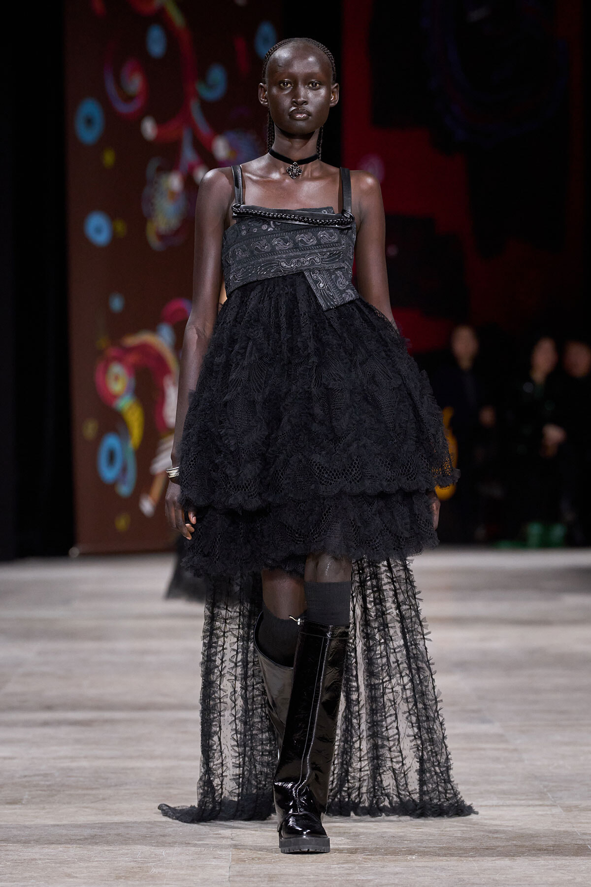 Black-clad female model walking a runway in a layered lace dress with a metallic corset-style bodice, choker, and tall shiny boots; colorful stage backdrop in background.