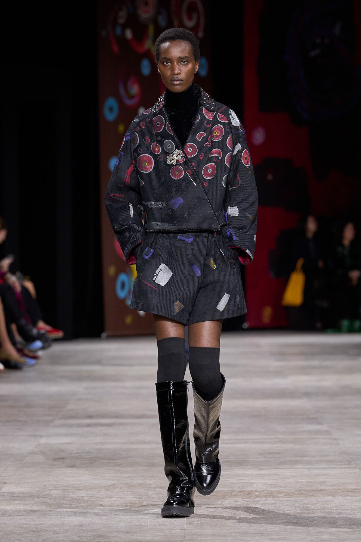 Model on a runway wearing a black patchwork jacket with circular red patterns, matching shorts, and tall boots.