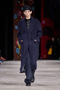 Male model walks the runway in a navy embroidered overcoat, black leather shirt, and a black turban-style hat.