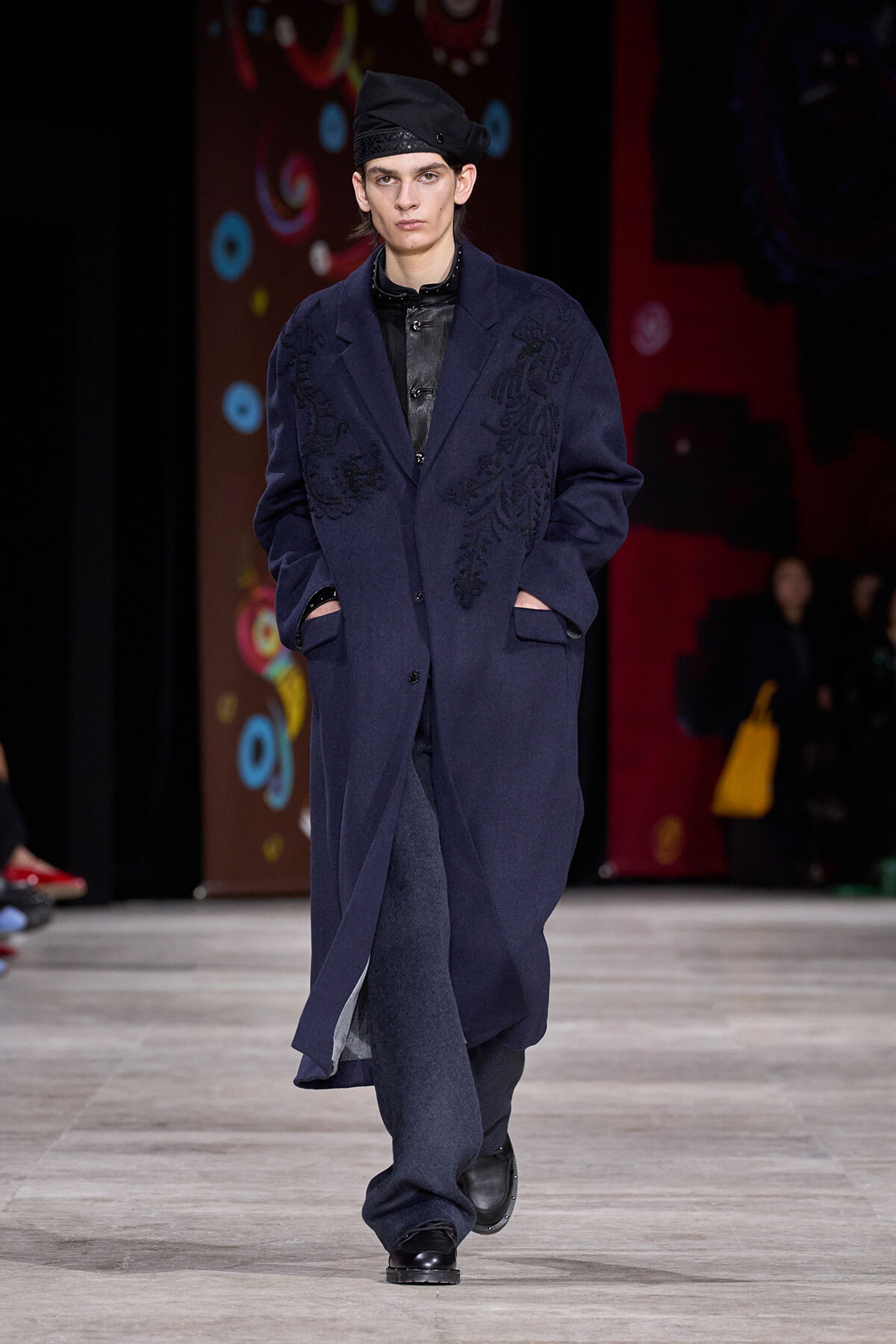 Male model walks the runway in a navy embroidered overcoat, black leather shirt, and a black turban-style hat.