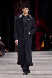 Male model walking a runway in a long black coat with fringe at the hem and a jeweled brooch on the lapel.