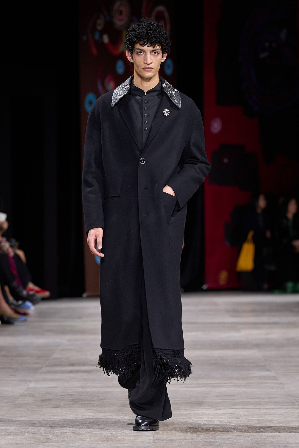 Male model walking a runway in a long black coat with fringe at the hem and a jeweled brooch on the lapel.