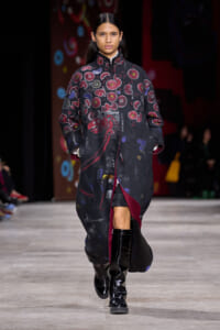 Model on a runway wearing a long black coat with red circular patterns and a burgundy interior, paired with knee-high black boots.