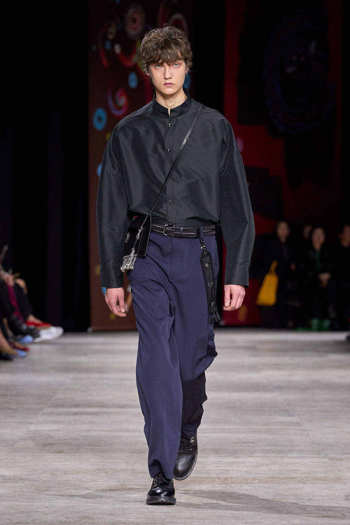 Male model walks a fashion runway in a dark button-up shirt, loose navy trousers, black belt, and a black crossbody bag with metallic charm.