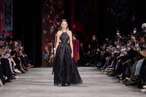 Runway moment: model in a black, sequined halter gown walking the catwalk as photographers crowd the sides.