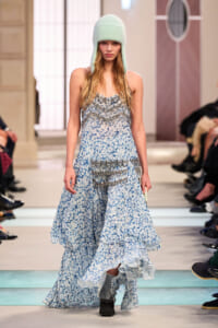 Model on a runway wearing a long blue floral gown with layered ruffles and a mint knit hat.