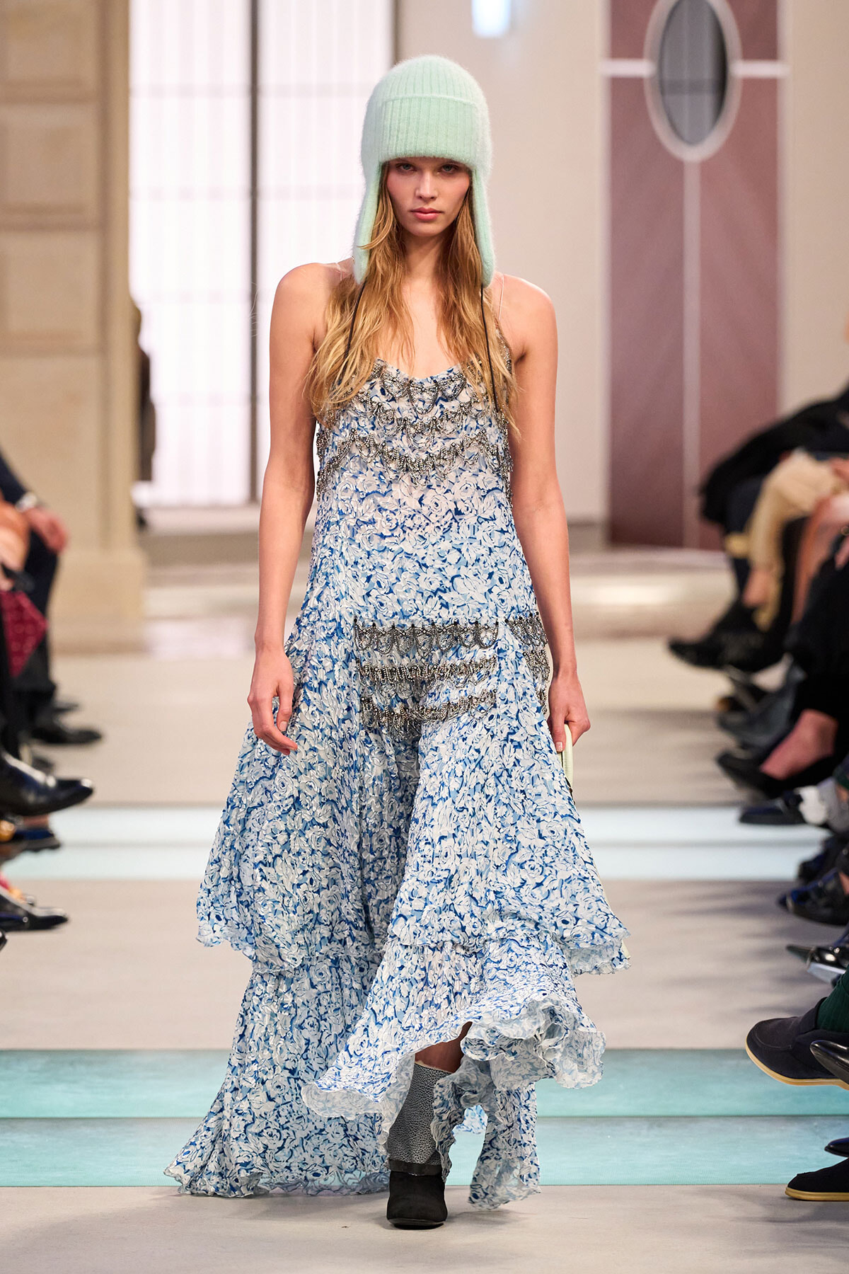 Model on a runway wearing a long blue floral gown with layered ruffles and a mint knit hat.