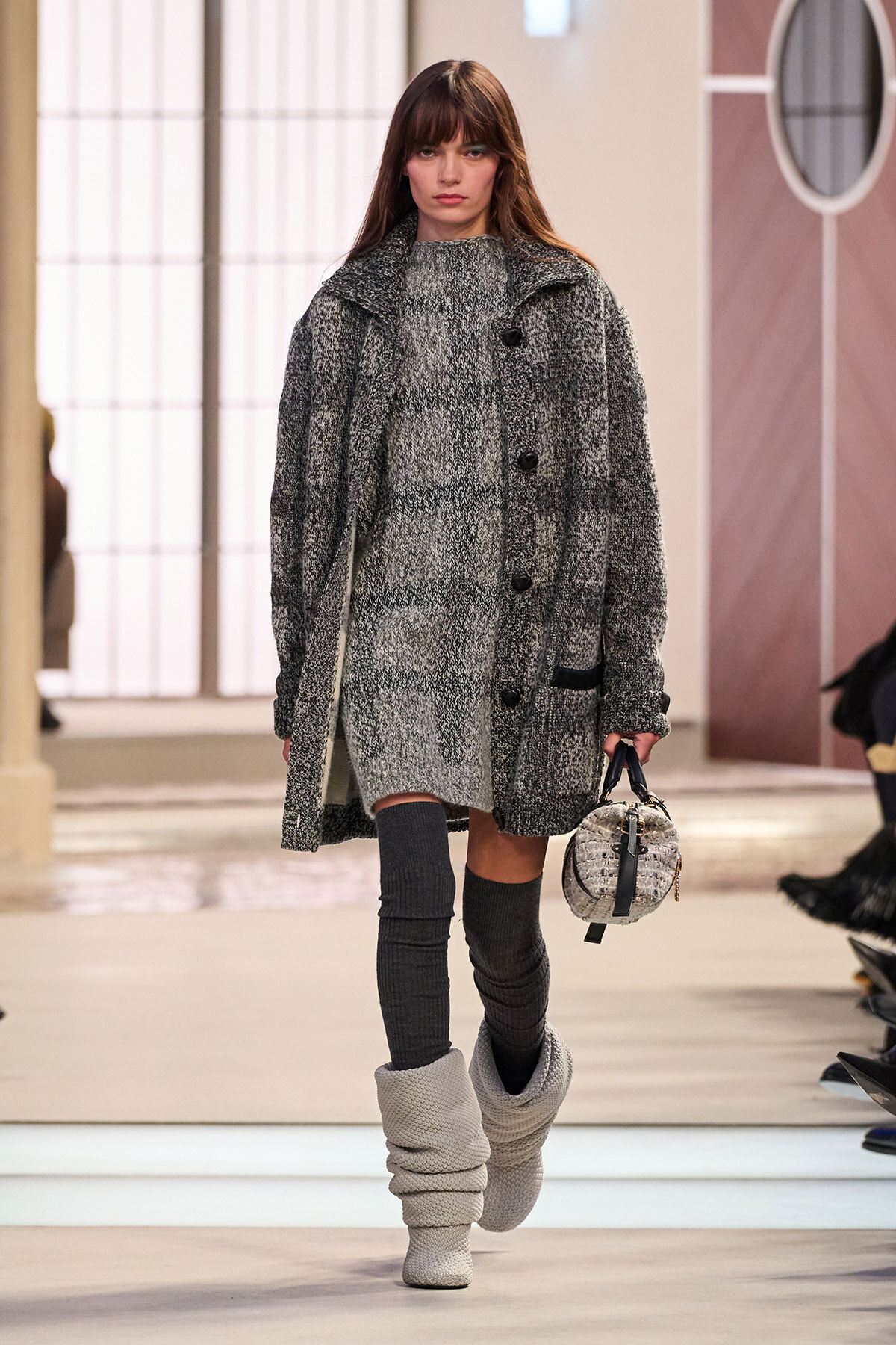 Runway model in a oversized gray tweed coat and matching knit dress, wearing chunky knit boots and carrying a small round tweed handbag.