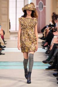 Model on a runway wearing a brown-yellow floral off-shoulder dress with a matching hat, gray knee-high socks, and dark blue boots.