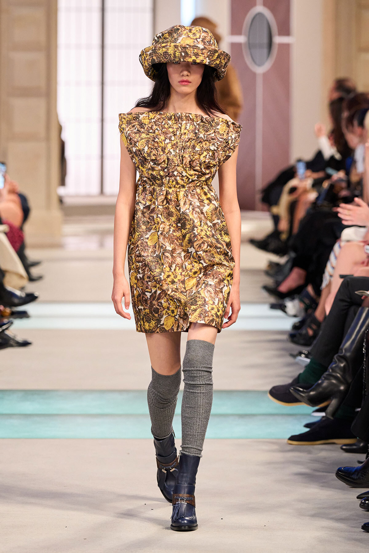 Model on a runway wearing a brown-yellow floral off-shoulder dress with a matching hat, gray knee-high socks, and dark blue boots.