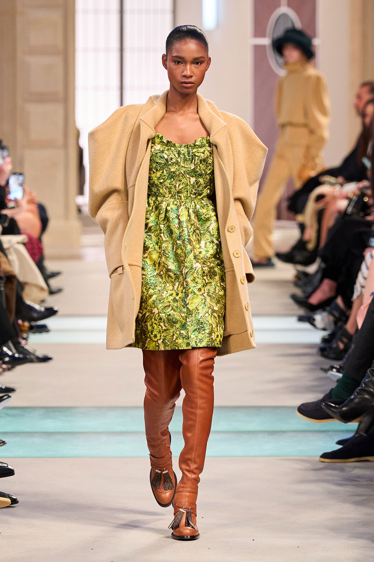 Model on a runway wearing a green botanical print dress, a beige oversized coat, and brown knee-high boots.