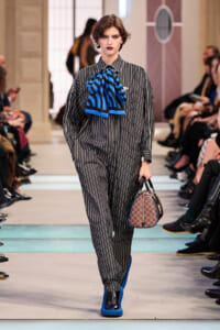 Model on a runway wearing a gray pinstripe jumpsuit with a bold blue striped necktie, cobalt boots, and a brown checkered handbag.