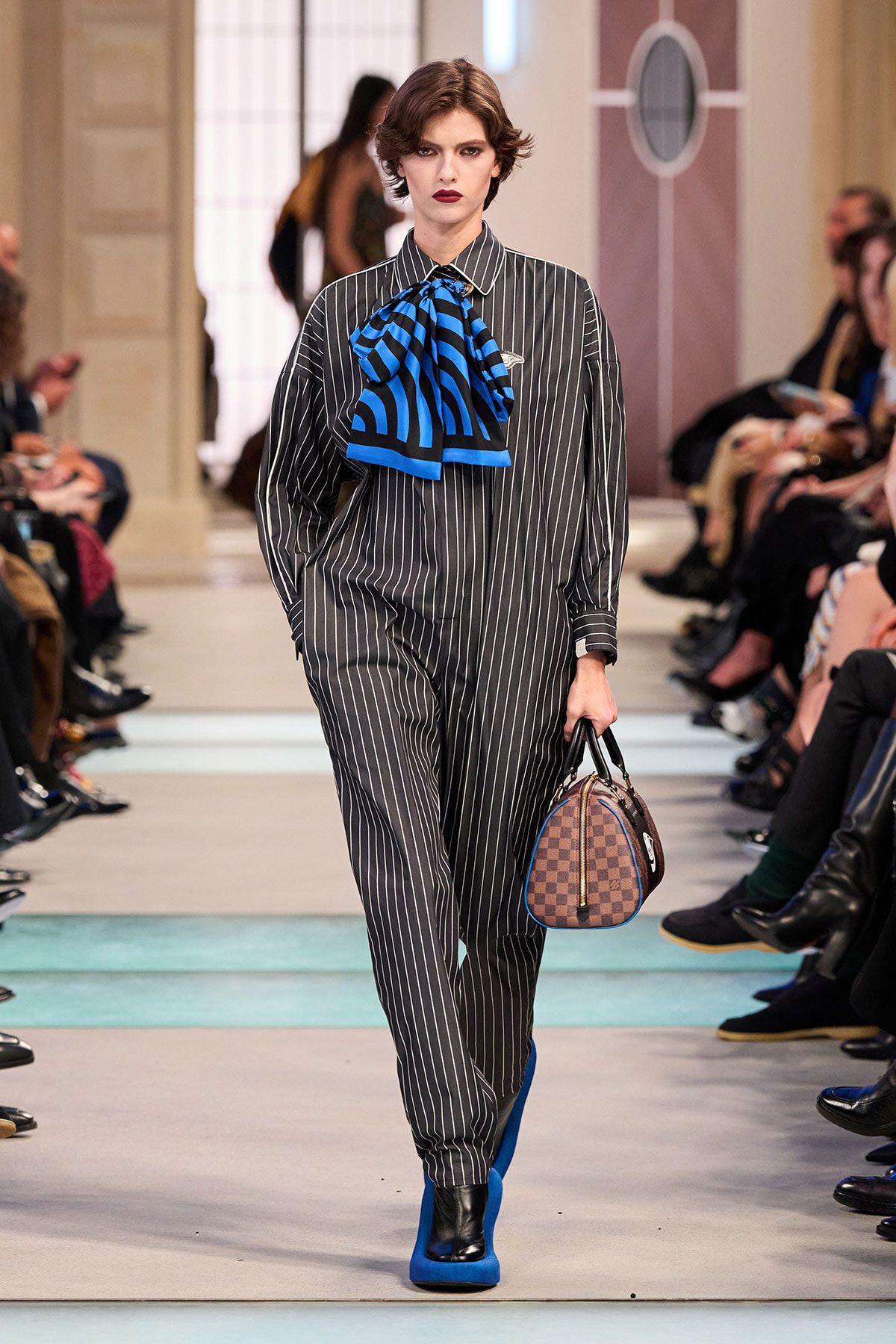 Model on a runway wearing a gray pinstripe jumpsuit with a bold blue striped necktie, cobalt boots, and a brown checkered handbag.