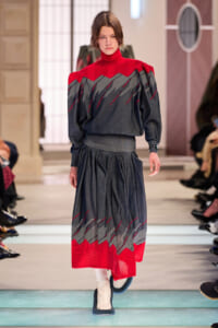 Model walking a runway in a red-and-gray knit dress with a high red turtleneck and sculpted shoulders.