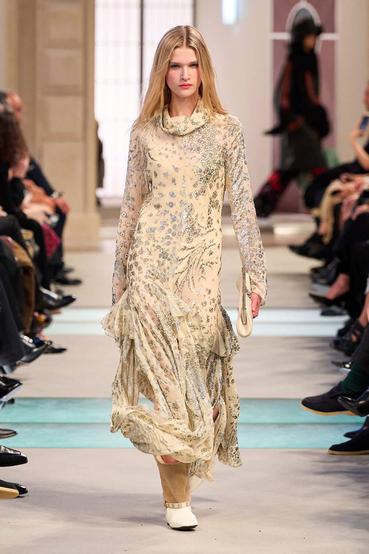 Model walks the runway in a cream, floral-embroidered gown with long sleeves and a layered, ruffled hem; audience on both sides observes.