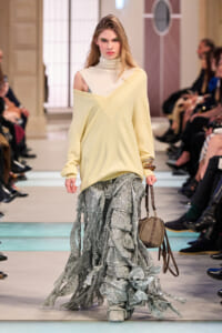 Model walking runway in an oversized pale yellow off-shoulder sweater over a one-shoulder top, paired with a layered silver-gray ruffled skirt and a small beige handbag.