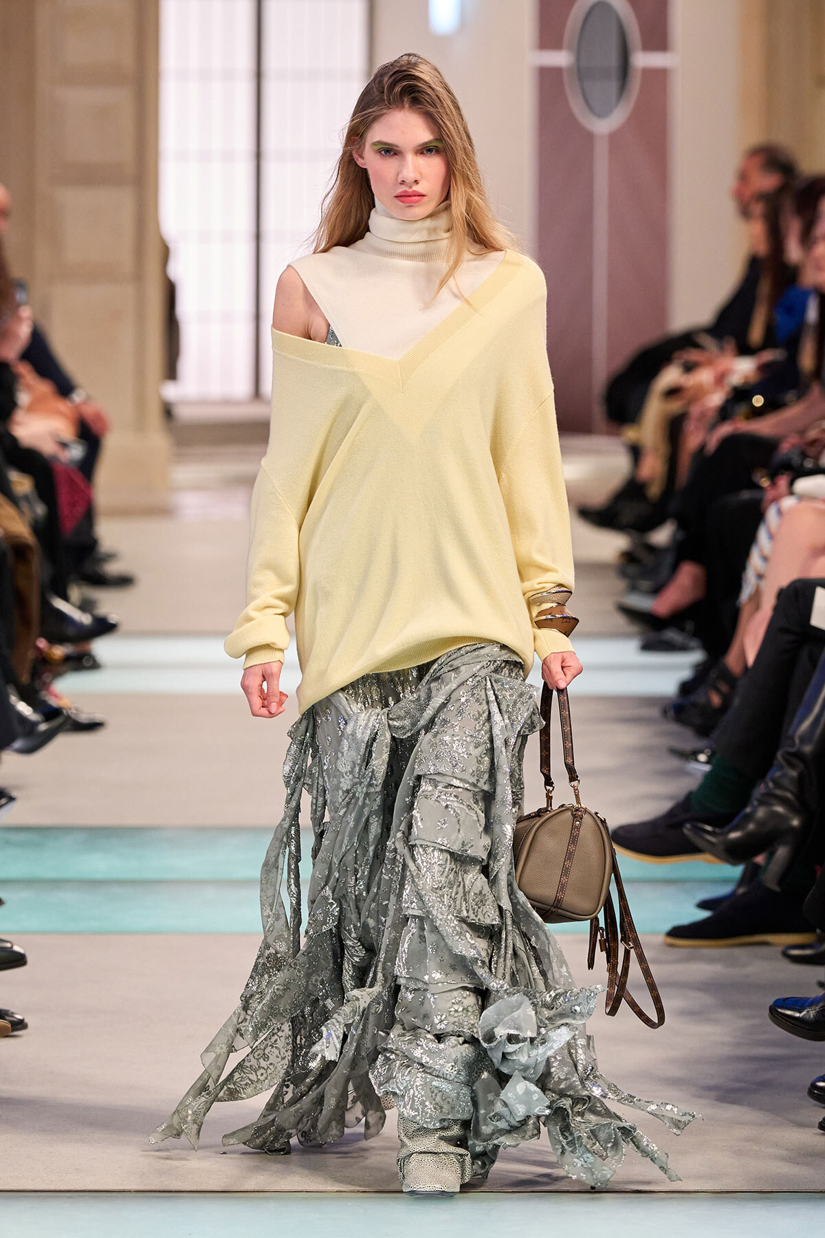 Model walking runway in an oversized pale yellow off-shoulder sweater over a one-shoulder top, paired with a layered silver-gray ruffled skirt and a small beige handbag.