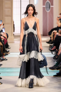 Model walks the runway in a navy slip dress with sheer lace panels and layered lace-trimmed skirt.