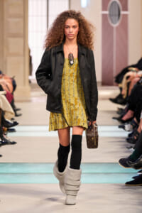 Model walking a runway in a yellow sequined mini dress with a black leather jacket, black knee-high socks, chunky white knit boots, and a small brown monogram handbag.