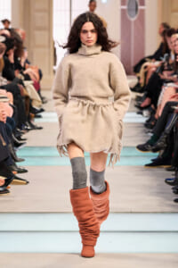 Model walks a beige oversized knit turtleneck dress with fringe, paired with gray knee socks and slouchy orange boots on a fashion runway.",