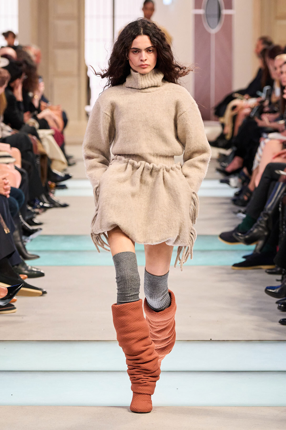 Model walks a beige oversized knit turtleneck dress with fringe, paired with gray knee socks and slouchy orange boots on a fashion runway.",