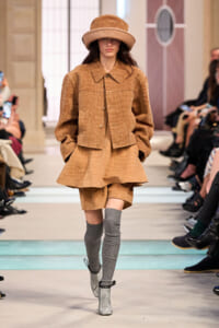 Model on a runway wearing a tan crocodile-textured two-piece suit with an oversized jacket, short skirt, gray knee-high socks, silver ankle boots, and a wide-brim hat.