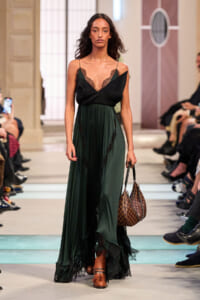 Model walks the runway in a dark emerald green gown with black lace trim and a brown checkered handbag in hand