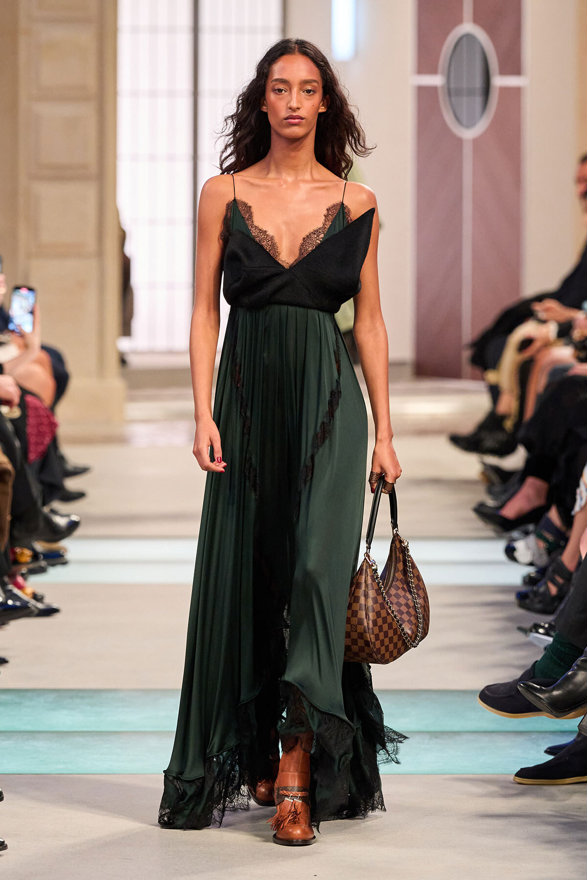 Model walks the runway in a dark emerald green gown with black lace trim and a brown checkered handbag in hand