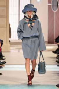 Model on a runway wearing a light blue belted dress, matching scarf and wide-brim hat, carrying a blue handbag; burgundy ankle boots.