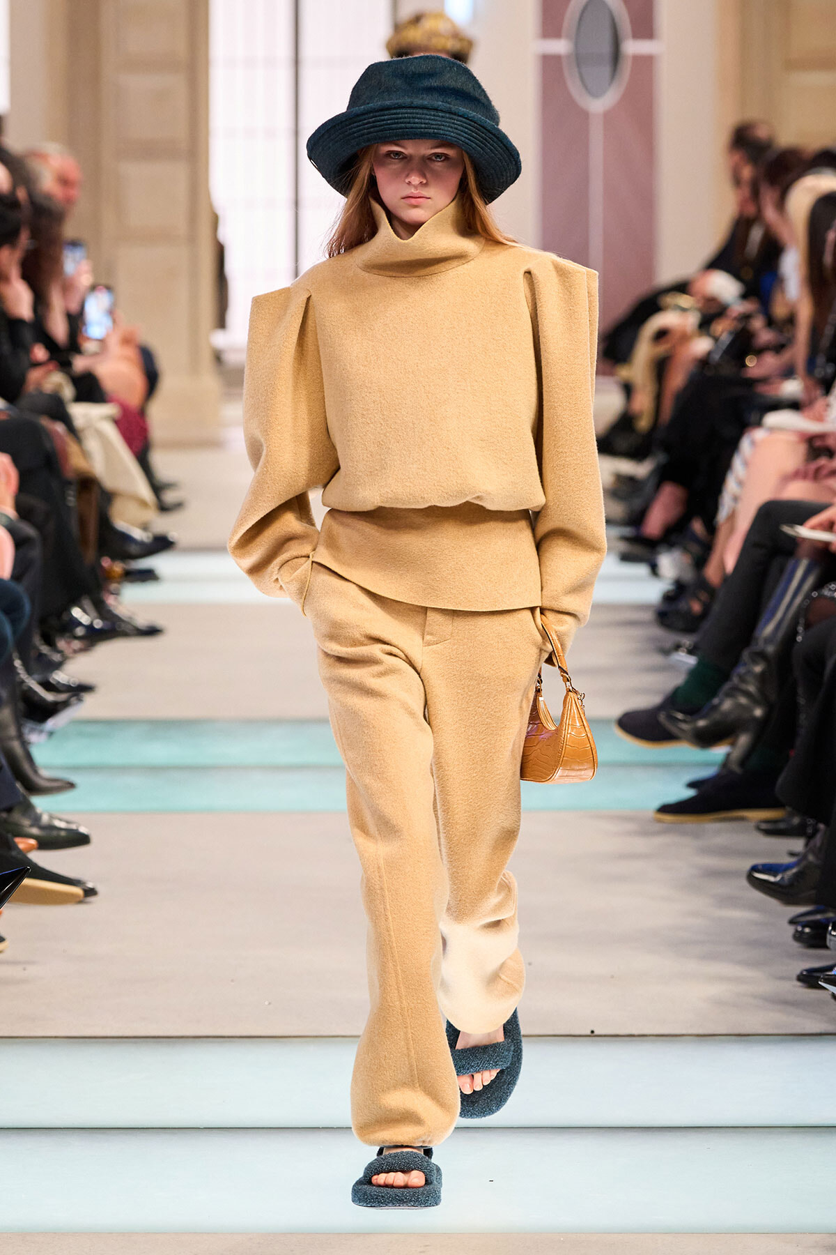 Runway model in a beige coordinated outfit with oversized shoulder details, wearing a dark blue hat and carrying a small tan handbag.