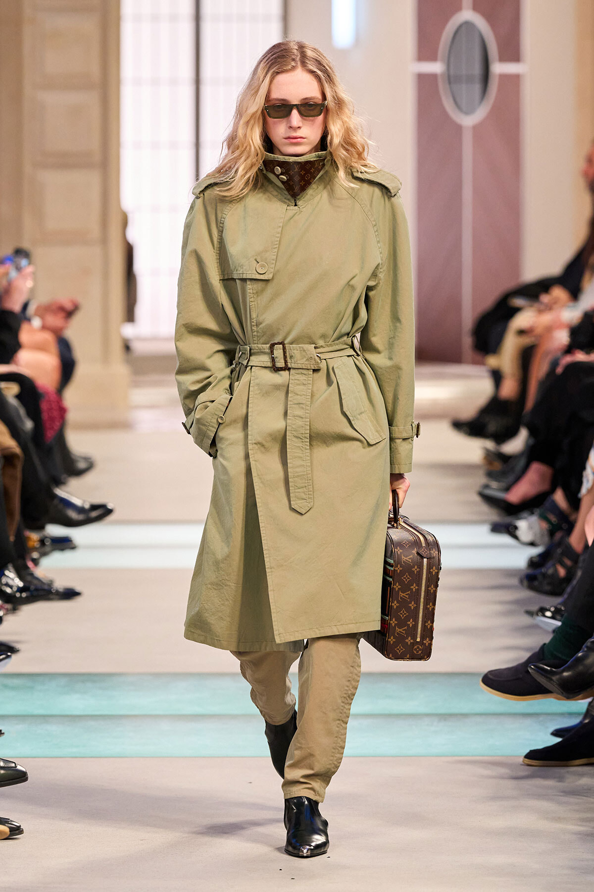 Model walks the runway in a olive trench coat with belt, sunglasses, and black ankle boots, carrying a brown Louis Vuitton suitcase on her right.