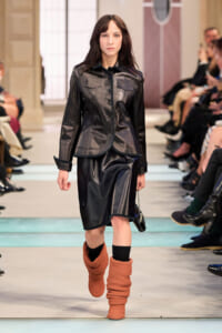Female model walking a fashion runway in a black leather jacket and matching skirt, paired with chunky orange boots; audience seated along the sides.