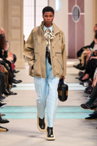 Model walking a runway in a beige utilitarian jacket, light blue striped pants, scarf, and a black handbag.
