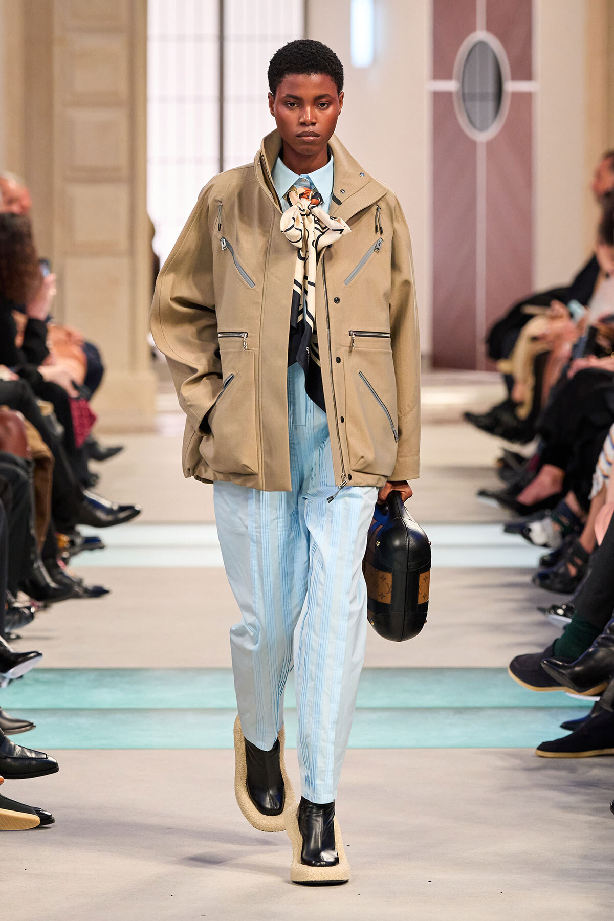 Model walking a runway in a beige utilitarian jacket, light blue striped pants, scarf, and a black handbag.