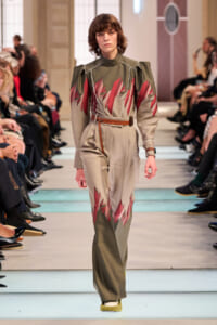 Model walks the runway in an olive-green and beige jumpsuit with bold red leaf-like patterns and a brown belt.