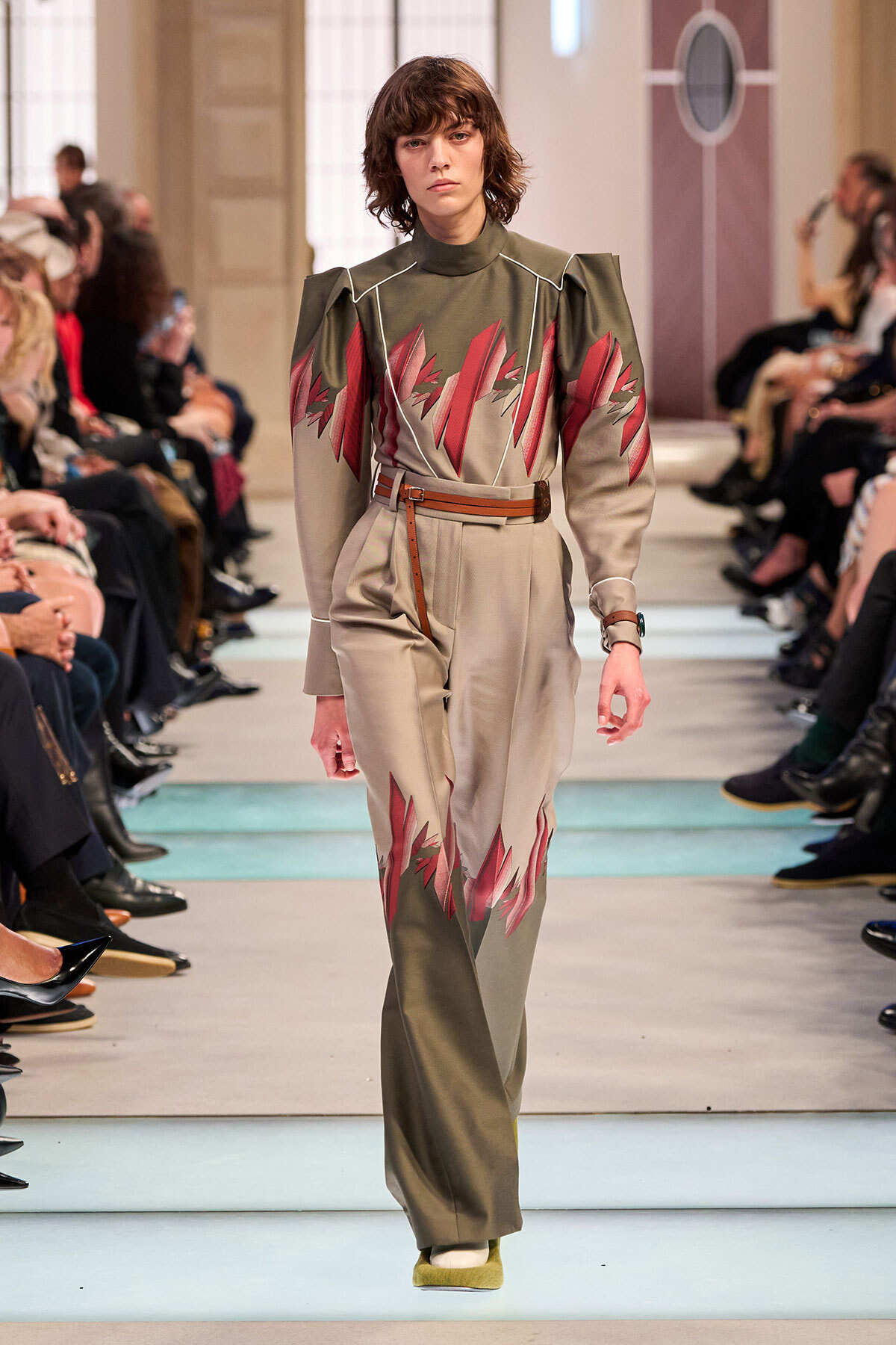 Model walks the runway in an olive-green and beige jumpsuit with bold red leaf-like patterns and a brown belt.