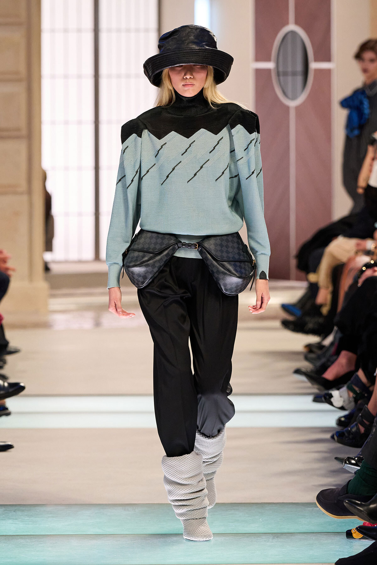 Runway look: pale blue sweater with a black yoke, black trousers, and a twin-pocket belt bag; model wears a wide-brim hat and light gray scrunched leg warmers.