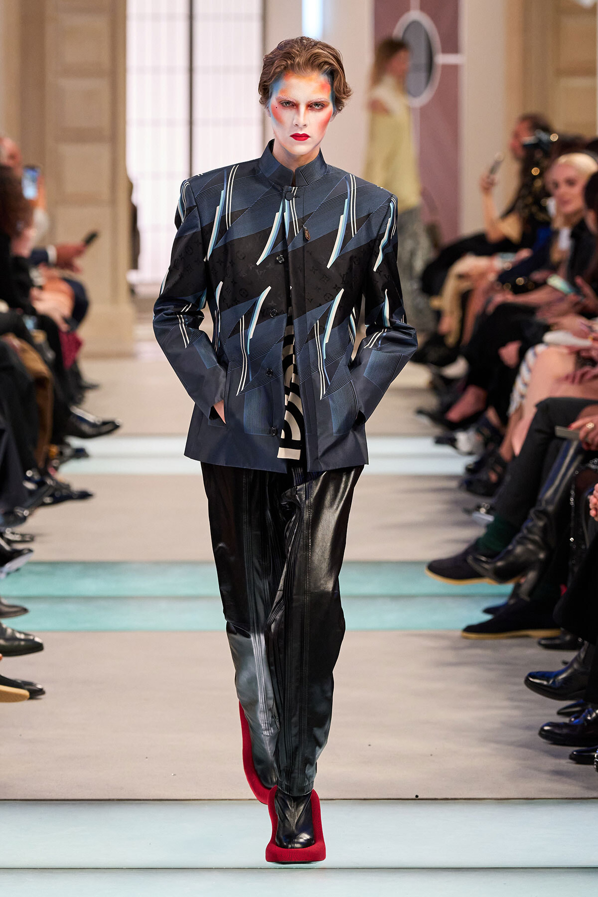 Model on runway wearing a structured geometric-pattern jacket in dark blue/black with blue lines, black leather pants, and bright red heeled boots; bold makeup visible behind.