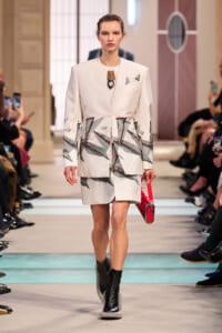 Female model on a fashion runway wearing a white blazer and matching shorts with a geometric gray and red print, paired with black ankle boots and a small red handbag. Audience members are seen watching and taking photos in the background.